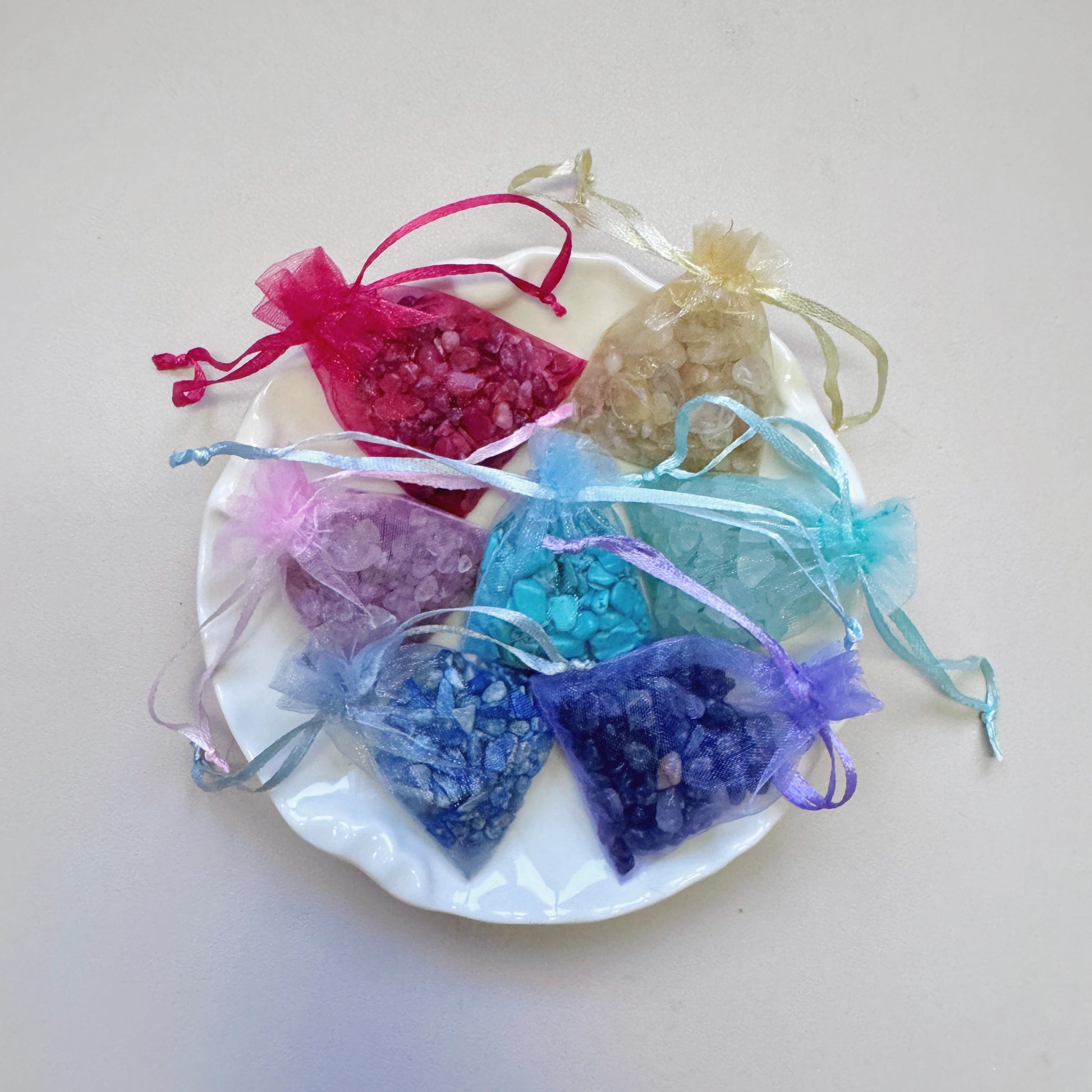 Seven organza bags with rainbow healing crystal chips—Nanhong Agate, Citrine, Strawberry Quartz, Green Fluorite, Turquoise, Sodalite, and Amethyst—displayed on a decorative plate.