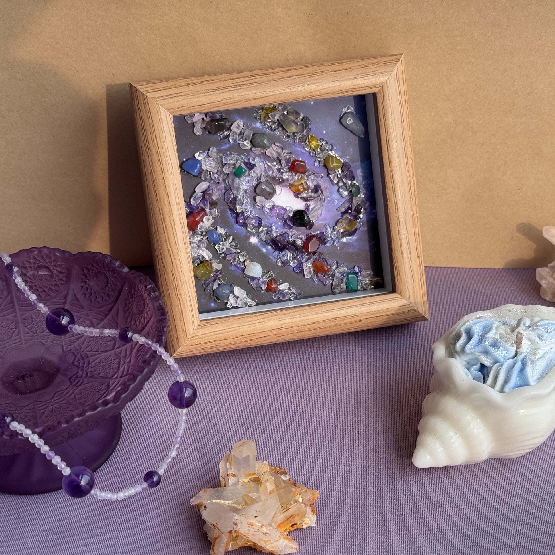 Detailed view of the framed Healing Crystal Spiral Painting Cosmic Art, showcasing the hand-placed natural gemstone galaxy. The artwork, in a dark wood frame, is surrounded by a spiritual room aesthetic, including an amethyst crystal bowl, a shell candle, and raw healing stone clusters for positive energy wall decor.