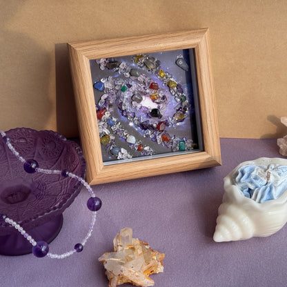 Detailed view of the framed Healing Crystal Spiral Painting Cosmic Art, showcasing the hand-placed natural gemstone galaxy. The artwork, in a dark wood frame, is surrounded by a spiritual room aesthetic, including an amethyst crystal bowl, a shell candle, and raw healing stone clusters for positive energy wall decor.
