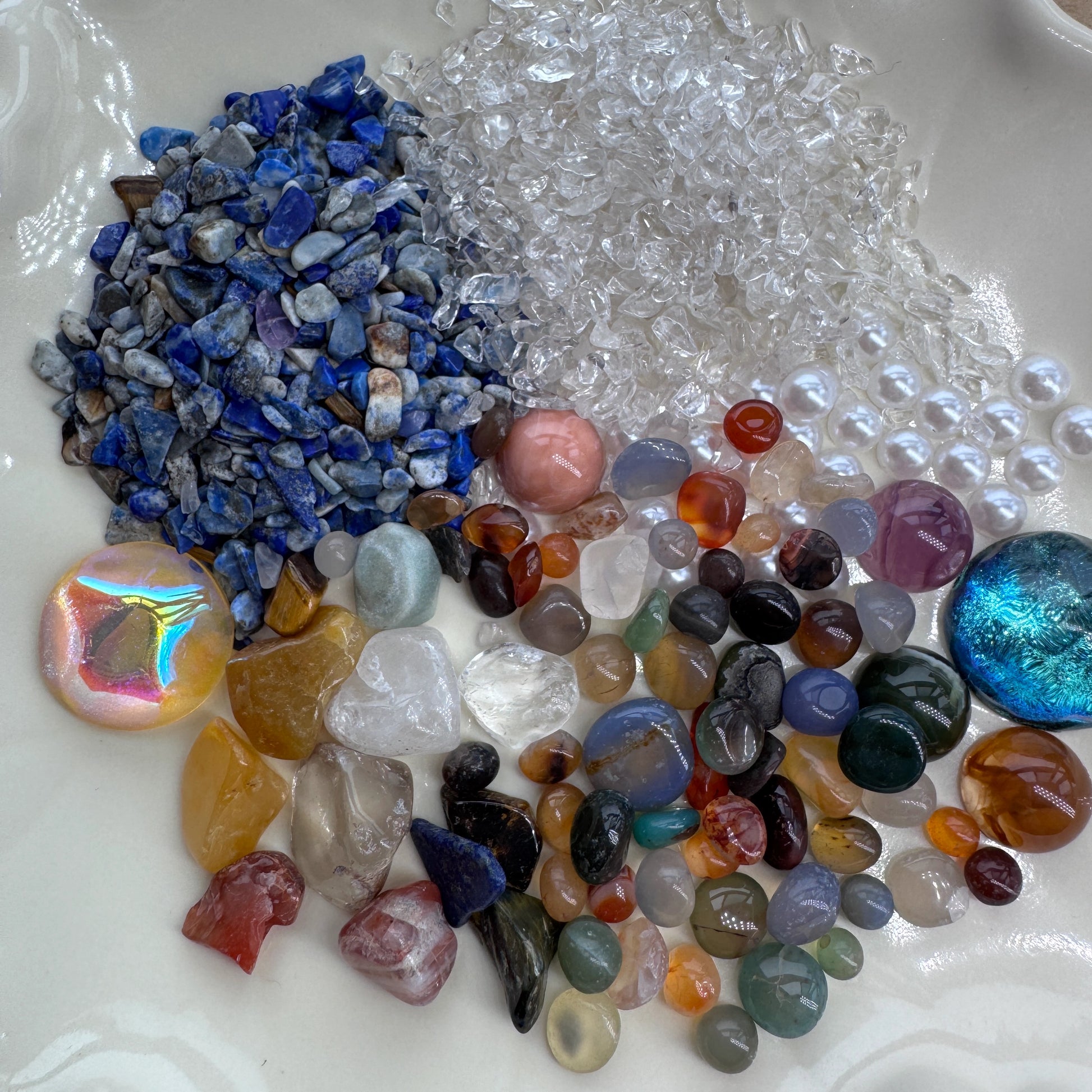 High-detail shot of various natural gemstone chips, pearls, and iridescent stones on a white plate.