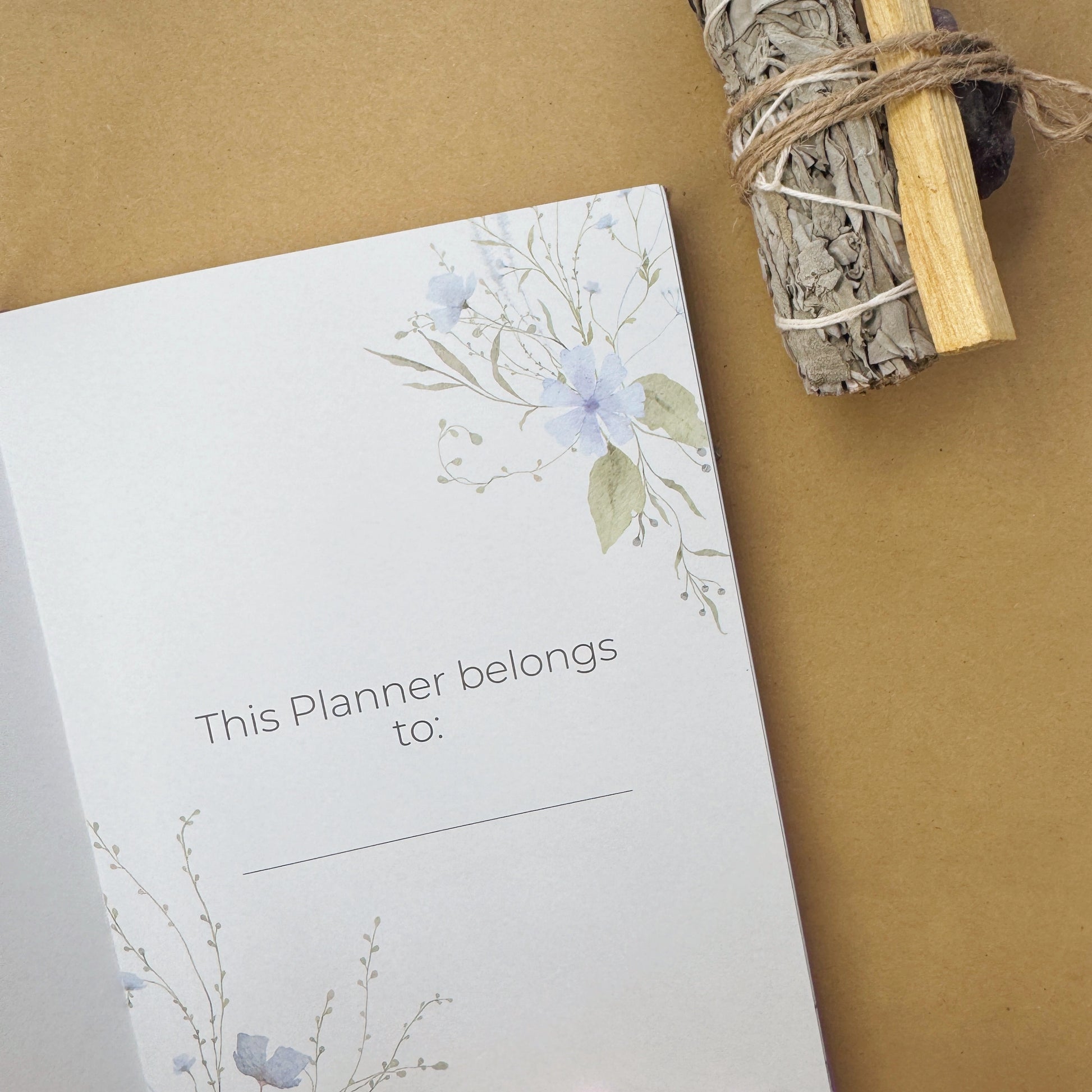 An open celestial mindfulness journal showing an intricate gold foil sun constellation title page, surrounded by raw amethyst white sage smudge sticks.
