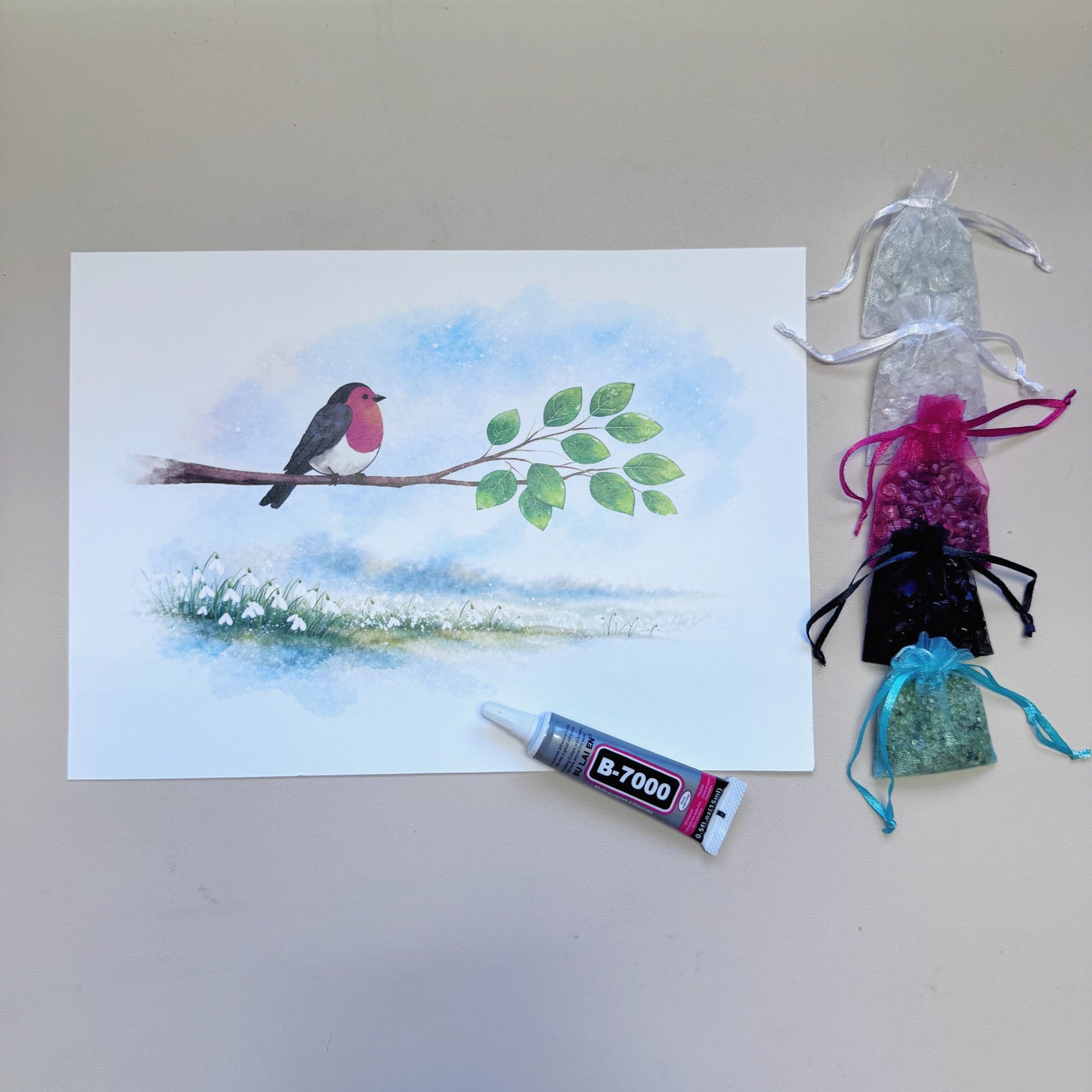 Robin and Snowdrop canvas art laid flat next to organized natural gemstone chips and a tube of B-7000 adhesive for DIY crystal mosaic craft.